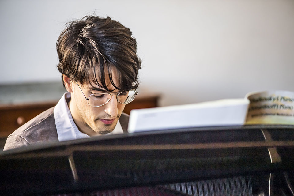 A male pianist, with dark hair and wearing glasses, looking down as he is playing the piano.